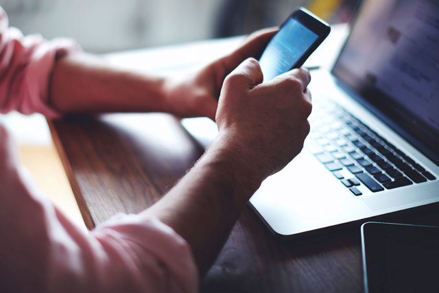 Mann bedient Smartphone vor einem Laptop sitzend